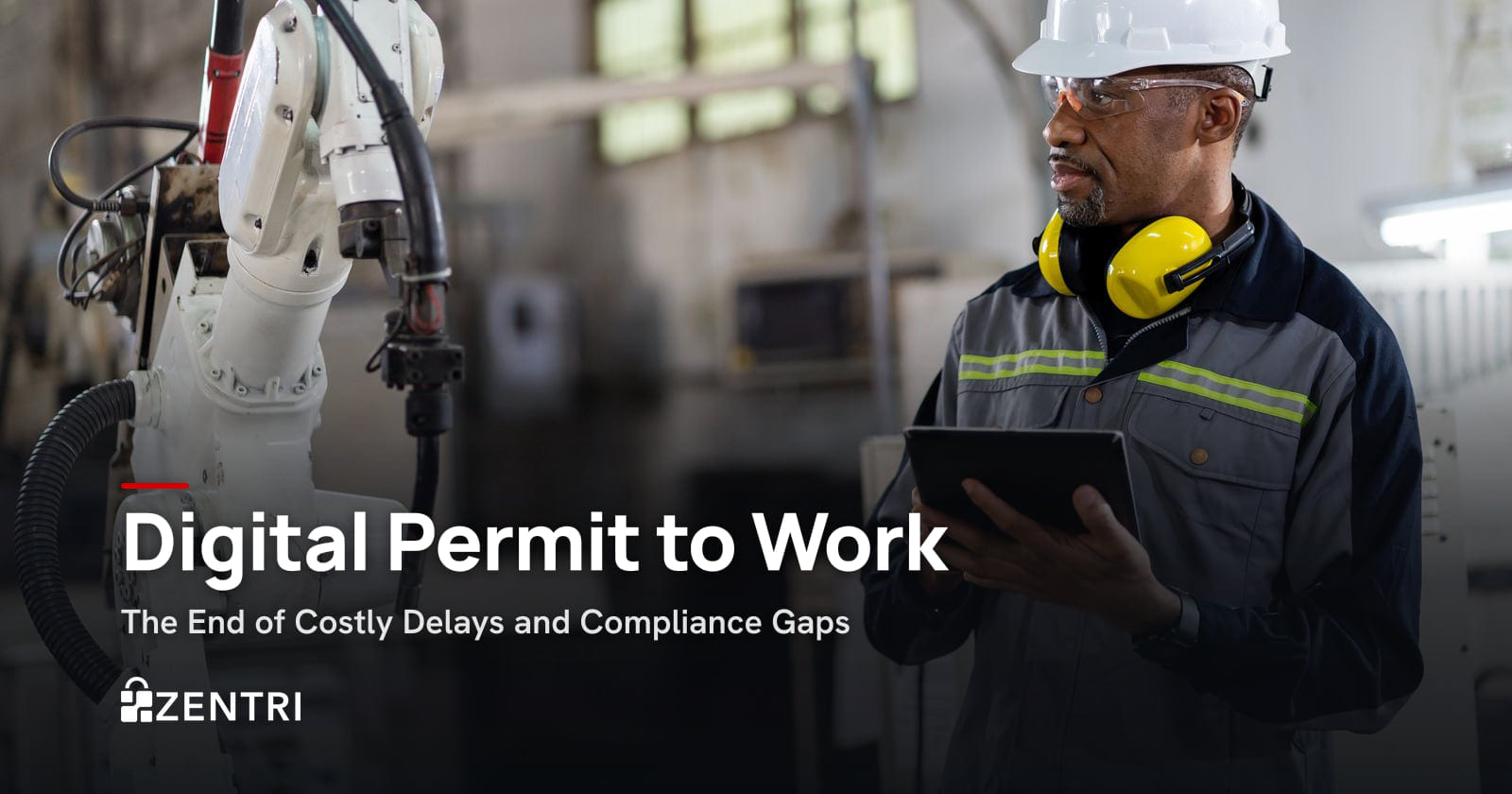 Engineer in a hard hat and ear defenders using a tablet beside a robotic arm